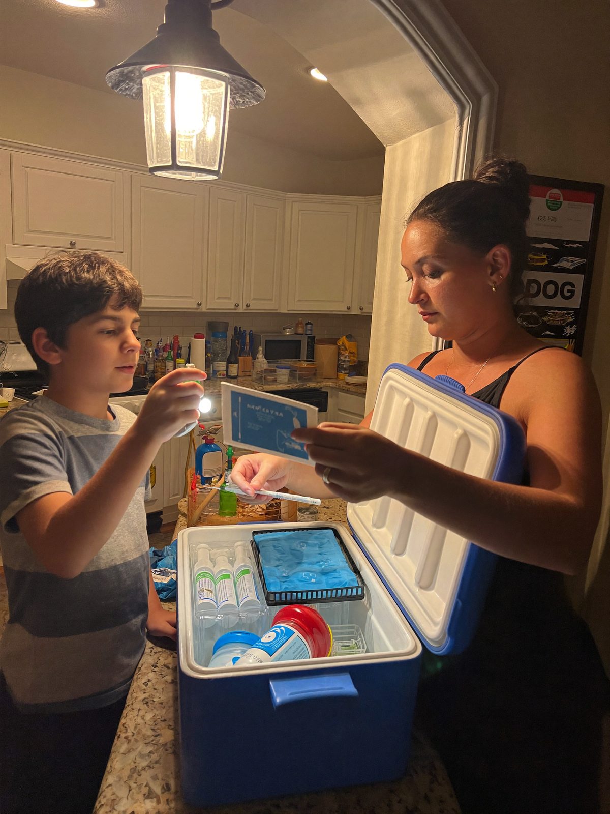 A mother checking an insulated cooler with medication and a thermometer while her son holds a flashlight