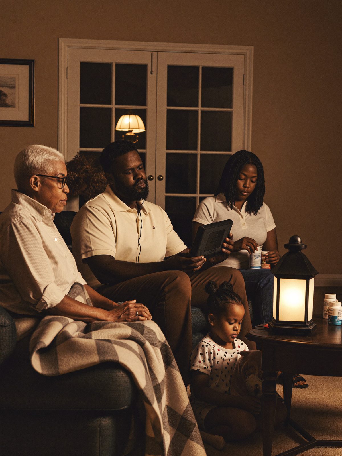 A multigenerational family coordinating by lantern light — grandmother seated, father with a radio, mother sorting medication, daughter holding the dog